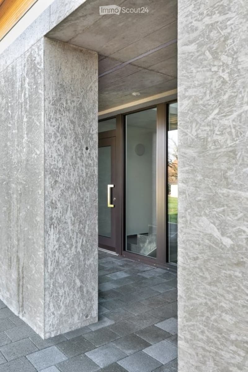 Modern building entrance with large concrete pillars, glass sliding doors, and a tiled pathway.