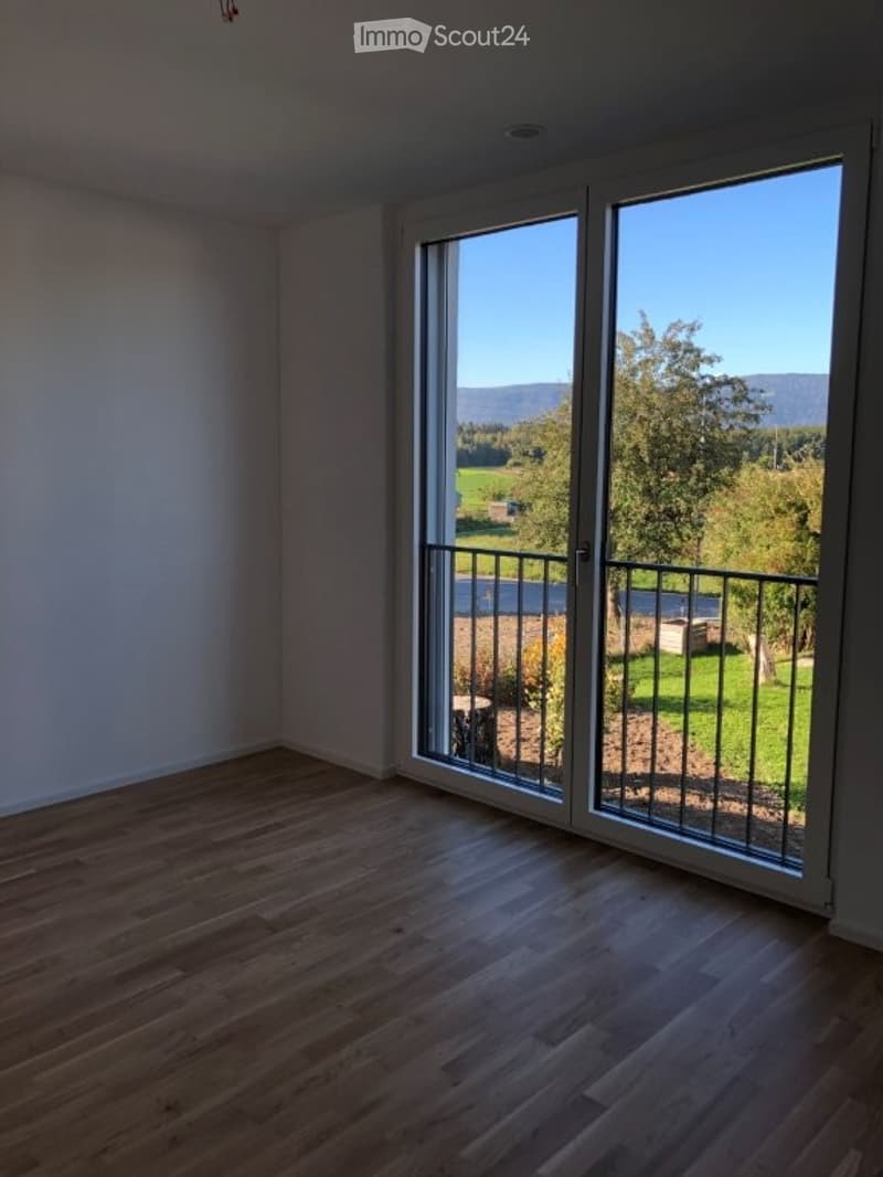 Empty room with wooden flooring and large sliding glass doors leading to a balcony with a scenic view of a green landscape and mountains.