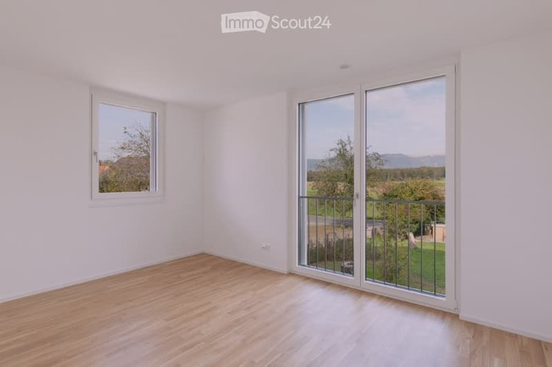 Empty room with light wooden flooring, white walls, and large windows, including a sliding glass door leading to a balcony.