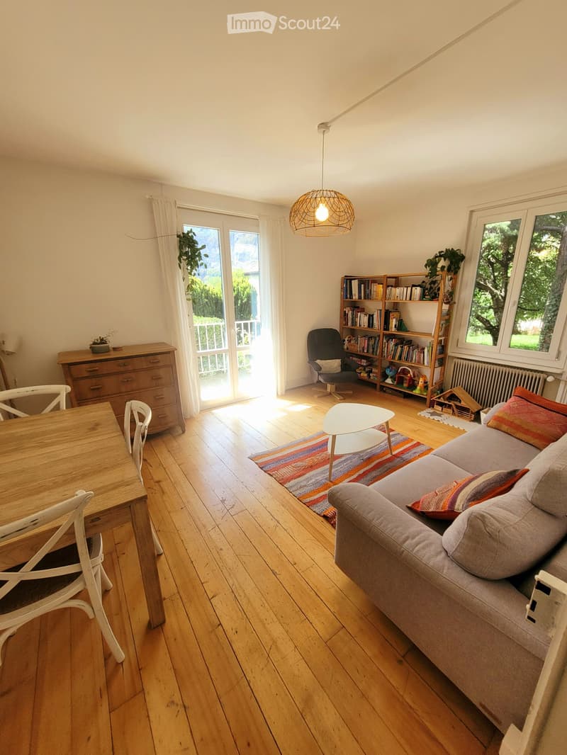 Bright living room with wooden flooring, large windows, and a balcony door. Features a grey sofa with colorful cushions, a white coffee table, a wooden dining table with chairs, a bookshelf, and a hanging pendant light.