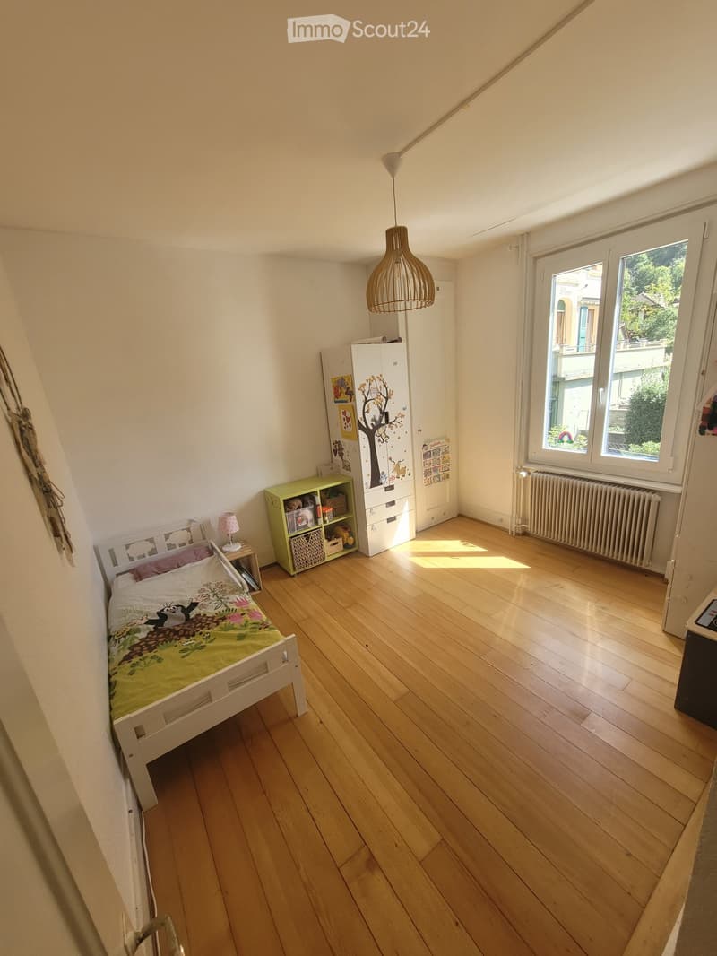 Bright bedroom with wooden flooring, a single bed with a colorful duvet, a nightstand with a lamp, a wardrobe, and a small shelf with storage boxes.