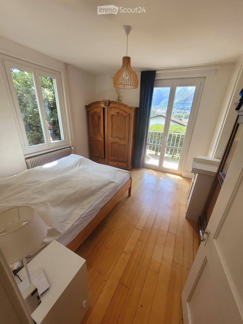 Bright bedroom with wooden flooring, large windows, and a scenic mountain view. Features a double bed, wardrobe, and pendant lighting.