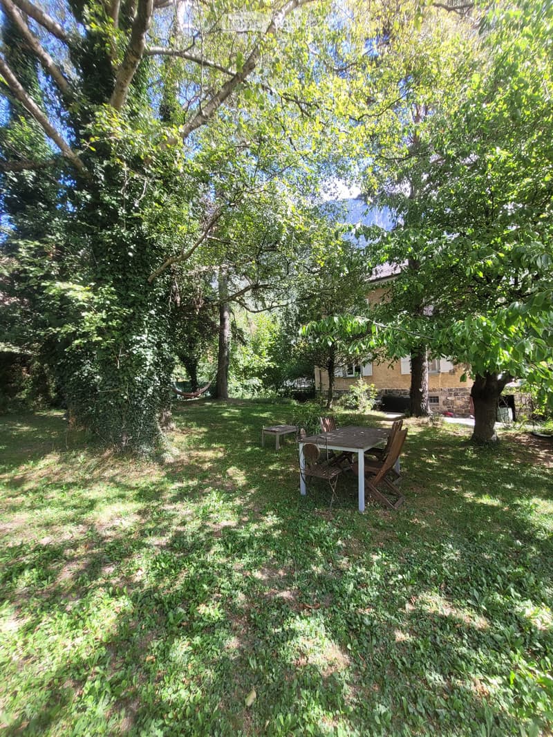 A lush garden with a table and chairs under a large tree, surrounded by greenery and a house in the background.