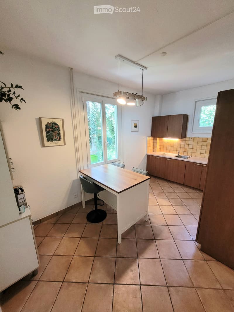 Bright kitchen with tiled floor, wooden table, chairs, and built-in cabinets. Large windows provide natural light.