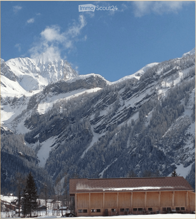 A wooden building with a brown roof and snow-covered ground, surrounded by snow-covered mountains and trees.