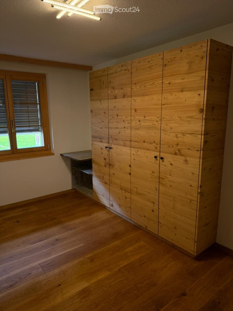 Empty room with wooden flooring, large wooden closet, window with blinds, and a small built-in shelf.