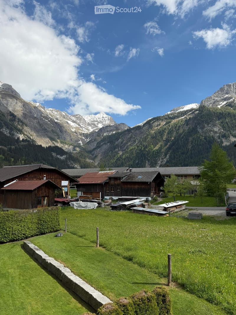 A picturesque alpine setting with wooden chalets, green lawn, and snow-capped mountains in the background.