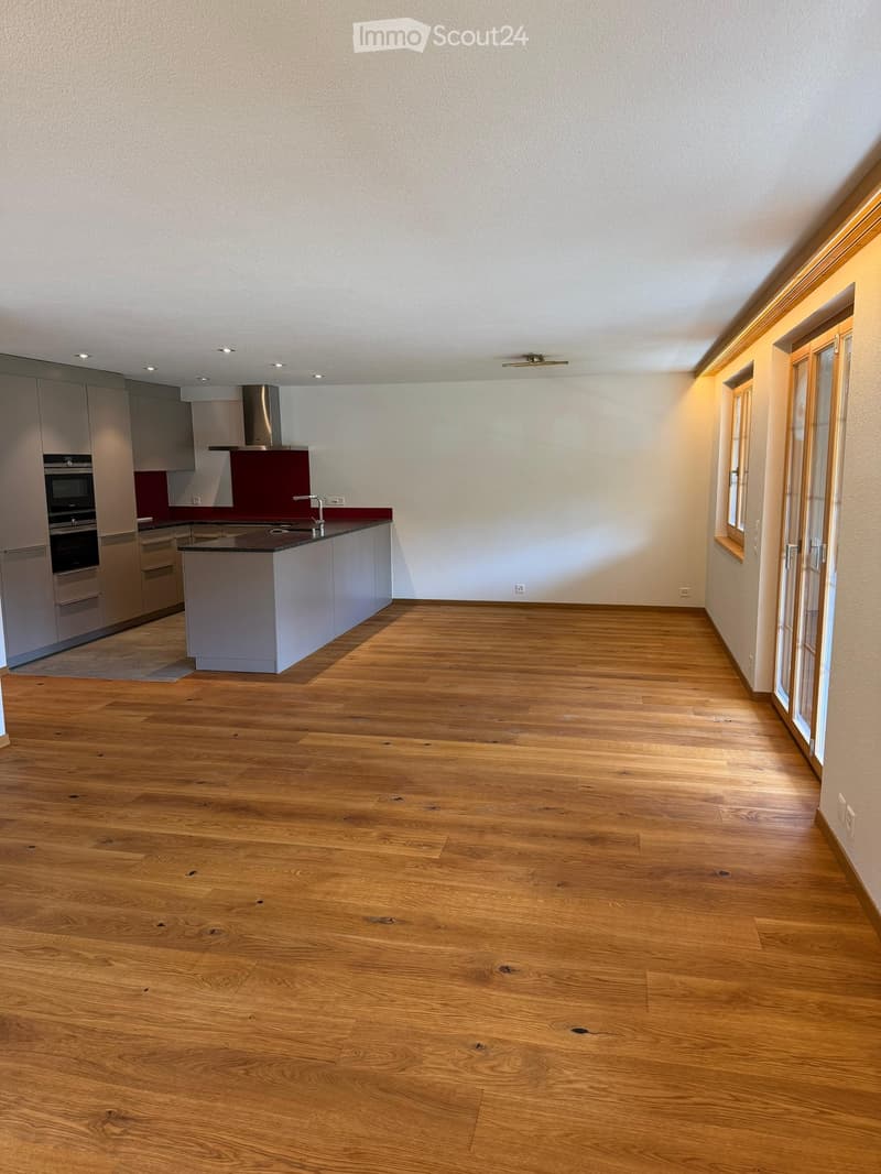 Empty room with light wooden flooring, white walls, and a modern kitchen area featuring grey cabinets, a stainless steel range hood, and a dark countertop.