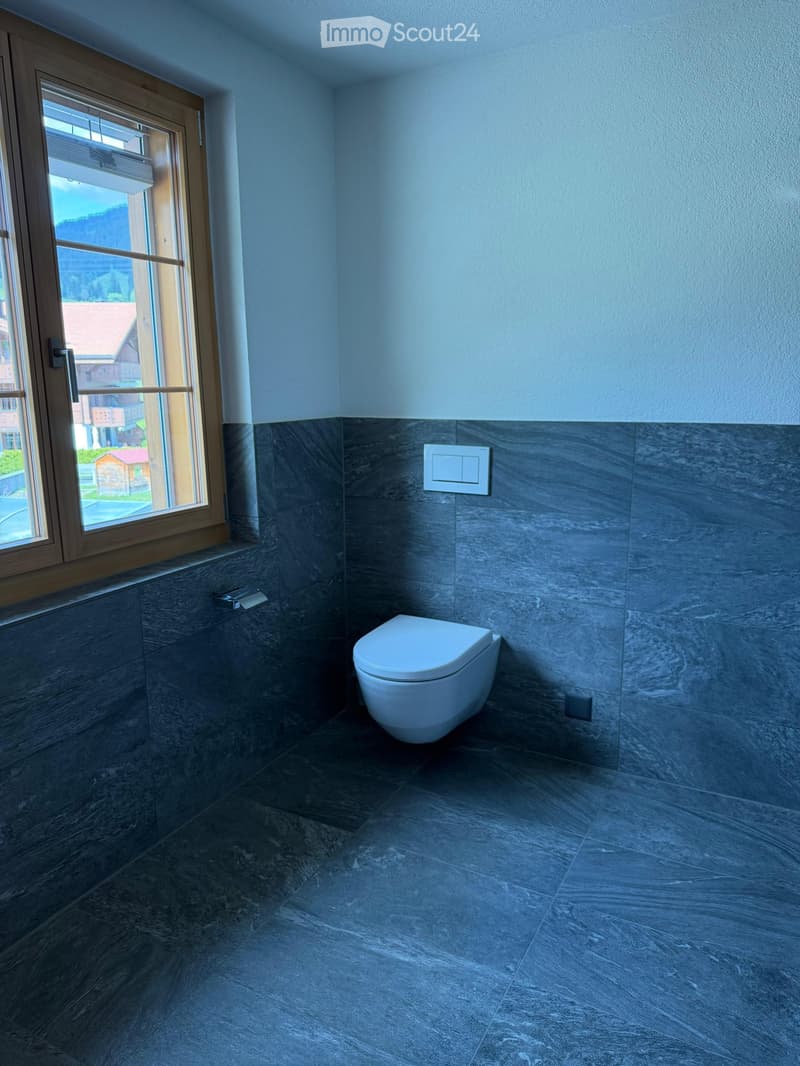 Modern bathroom with dark grey marble tiles, white toilet, and large window with wooden frame.