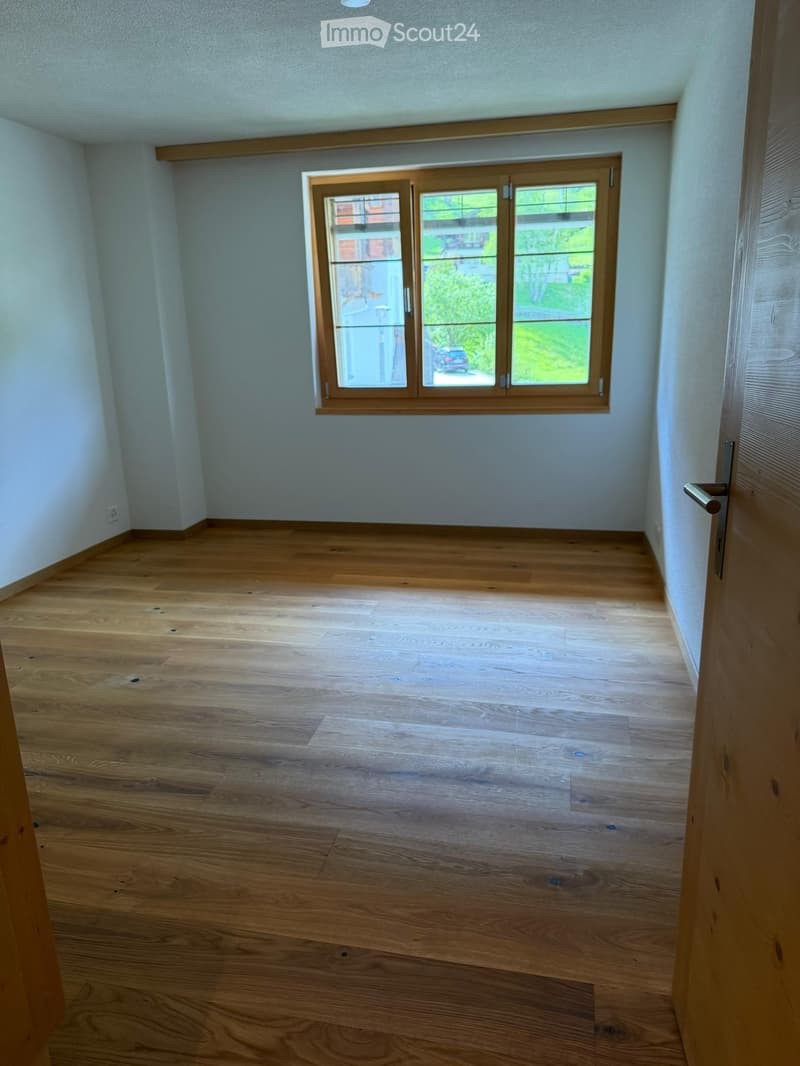 Empty room with light wooden flooring, white walls, and a large window with wooden frame showing a view of the garden and street.