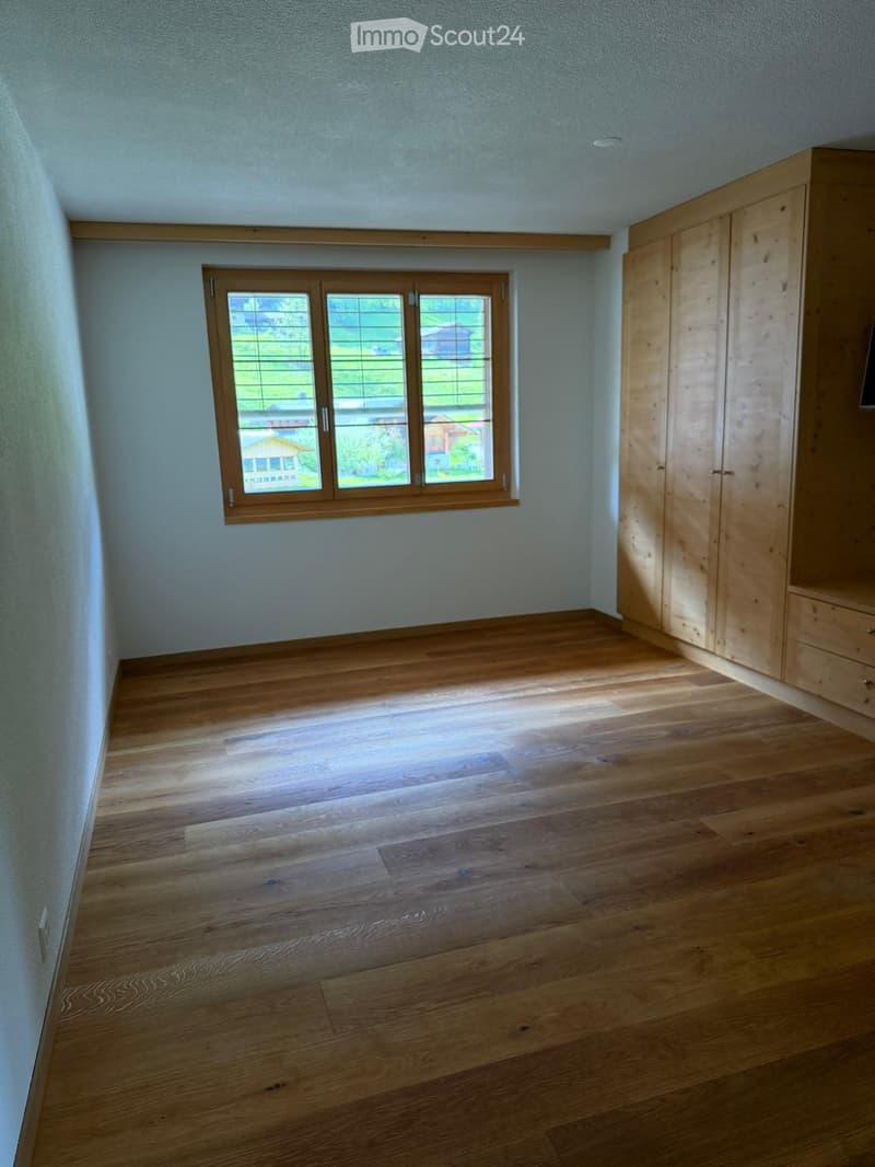 Empty room with wooden flooring, light-colored walls, and a large window with wooden frame and blinds. A built-in wooden wardrobe is visible on the right side.