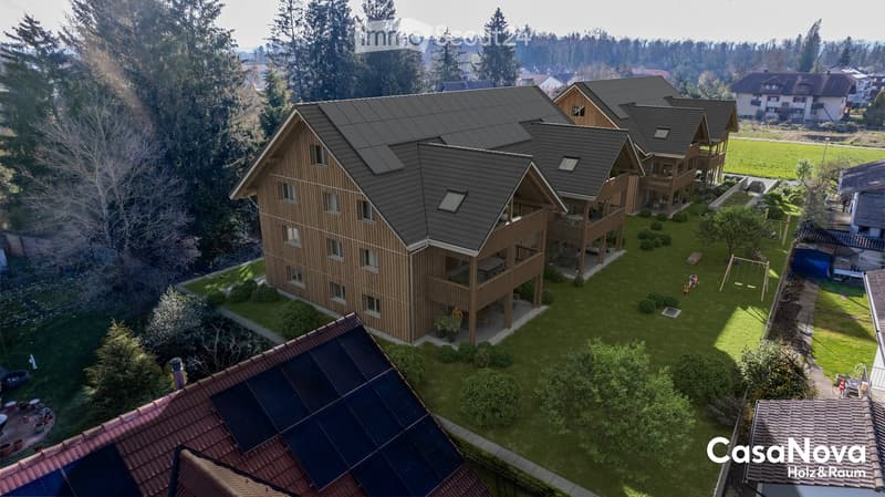 Aerial view of 4 identical detached houses, each with solar panels on the roof, wooden exteriors, multiple windows, balconies, surrounded by trees and lawns, green field in the background.