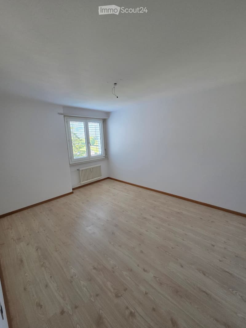 Empty room with light-colored wooden flooring, white walls, and a single window with blinds. A radiator is visible below the window.