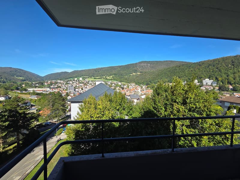 Balcony overlooking a town with houses, trees, and distant mountains under a clear blue sky.