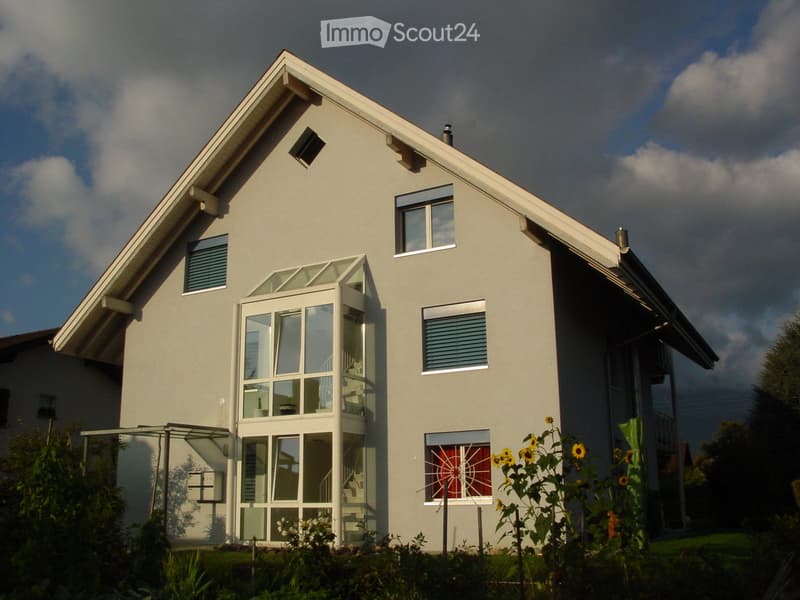 Two-story house with light gray exterior, large windows, and a modern design. Features include a prominent entrance with glass doors, a small balcony, and decorative sunflowers in the front yard.