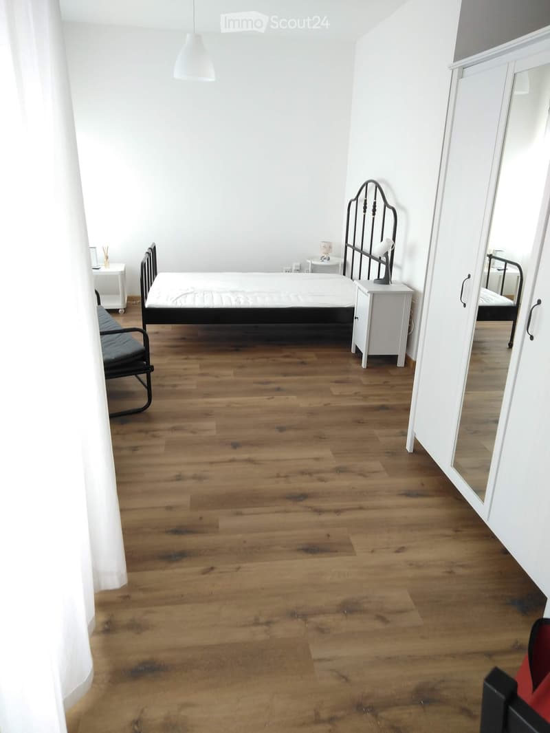 Modern bedroom with wooden flooring, a bed with a black metal frame, a nightstand, a chair, and a large mirror on the wall.