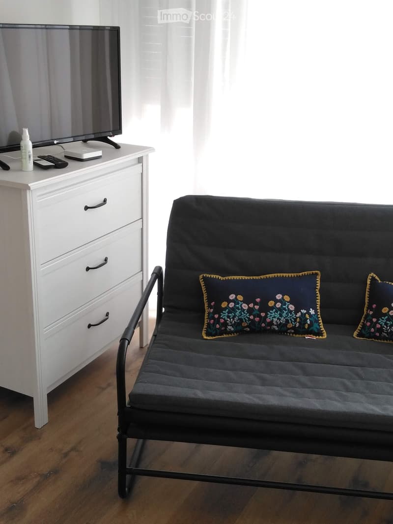 A small, modern living space with a black sofa, decorative pillows, a white dresser, and a television on top.