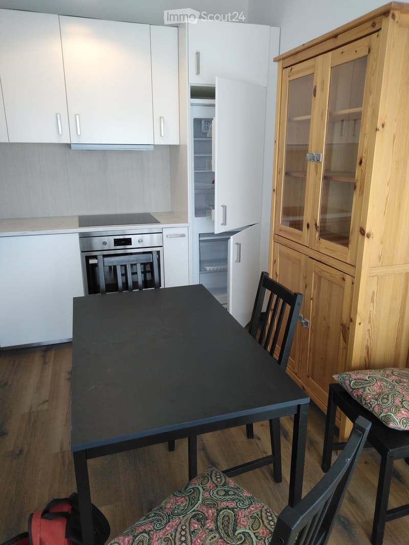 Modern kitchen with white cabinets, a black dining table, and two chairs with patterned cushions. An oven and refrigerator are visible, along with a wooden display cabinet.