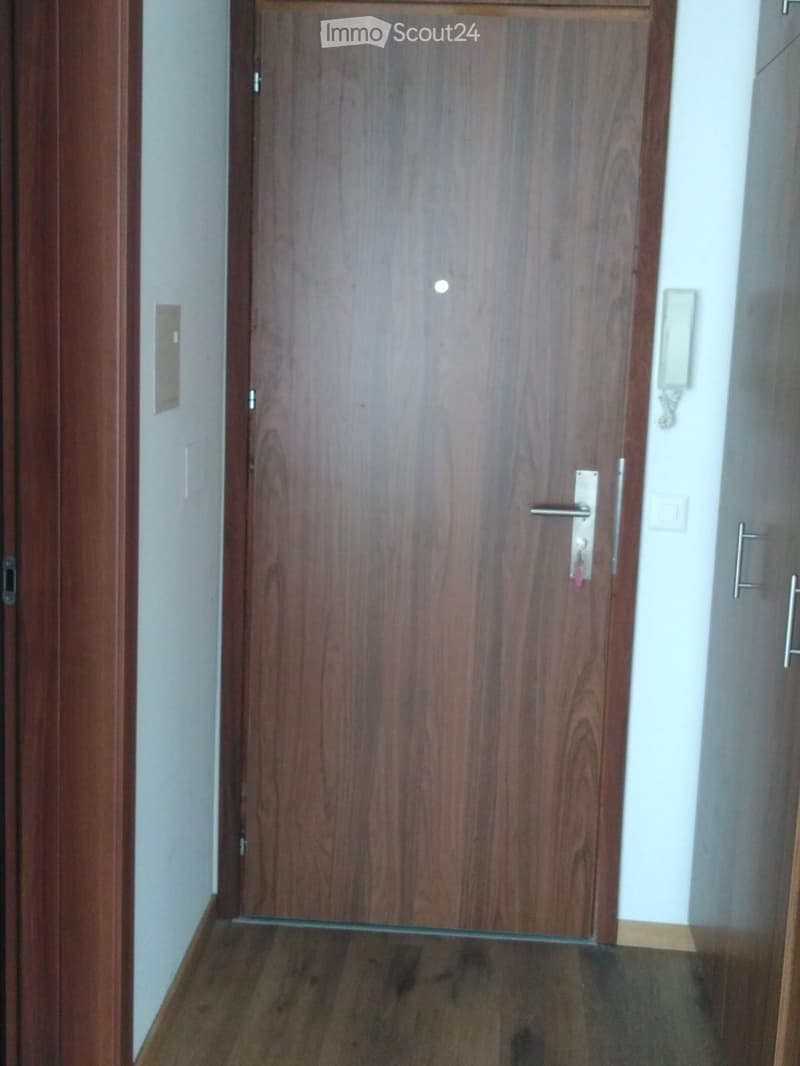 A wooden door with a silver handle and lock is closed, set in a wooden-framed doorway. The surrounding walls are light-colored, and the floor is made of dark wood.