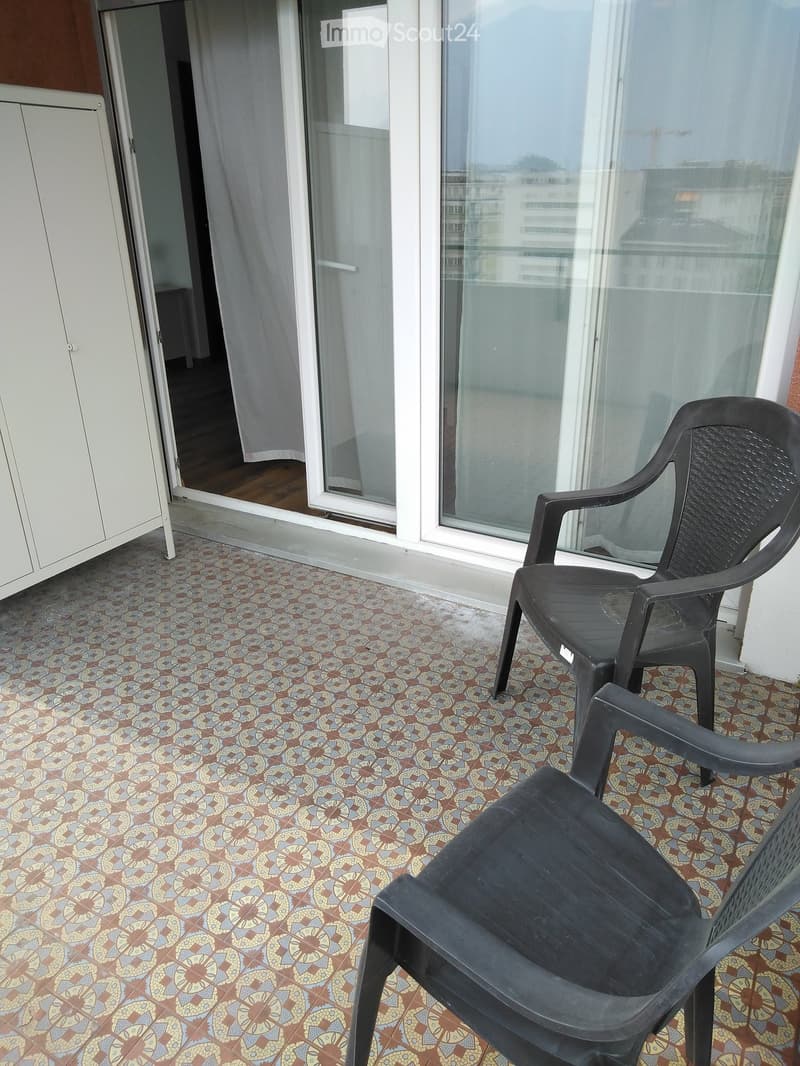 Balcony with patterned tile floor, two black plastic chairs, sliding glass door leading to interior, white cabinet visible on left.