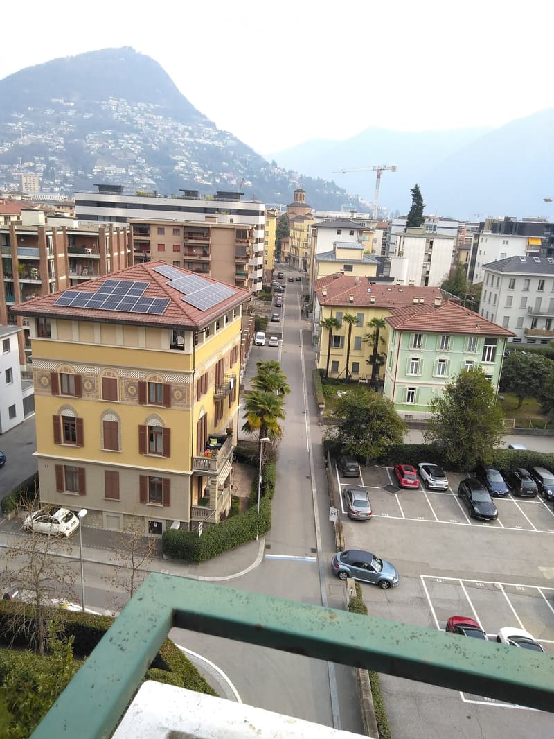 View from a balcony showing a street lined with buildings, parked cars, and a distant mountain range.