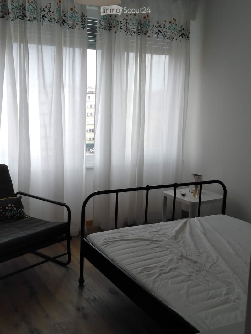 A small bedroom with a single bed, a chair, a small table, and a lamp. The room has wooden flooring and large windows with white curtains.