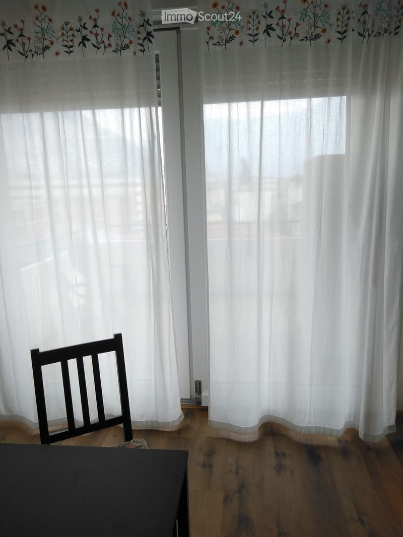 A partially furnished room with wooden flooring, white curtains, a black chair, and a black table.