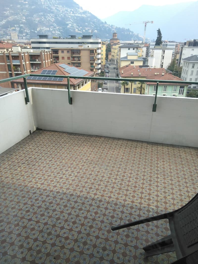 Balcony with patterned tile flooring, white walls, metal railing, and a scenic city view with buildings and mountains in the background.