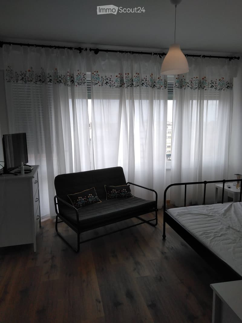 A small, well-lit room with wooden flooring, a sofa, a bed, a small table, and a hanging lamp. The room features large windows with white curtains and a television on a stand.