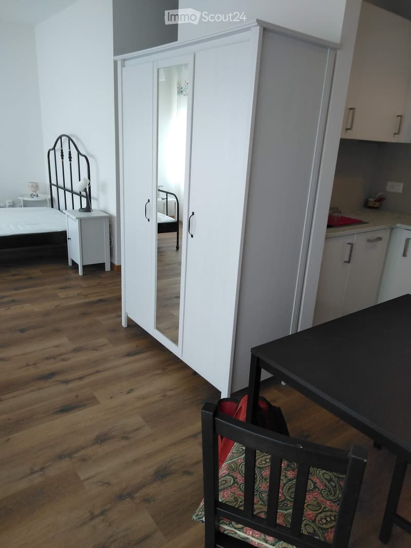 A furnished room with a wooden floor, a bed with a decorative headboard, a nightstand with a lamp, a wardrobe with mirrored doors, a small table, and a patterned chair.