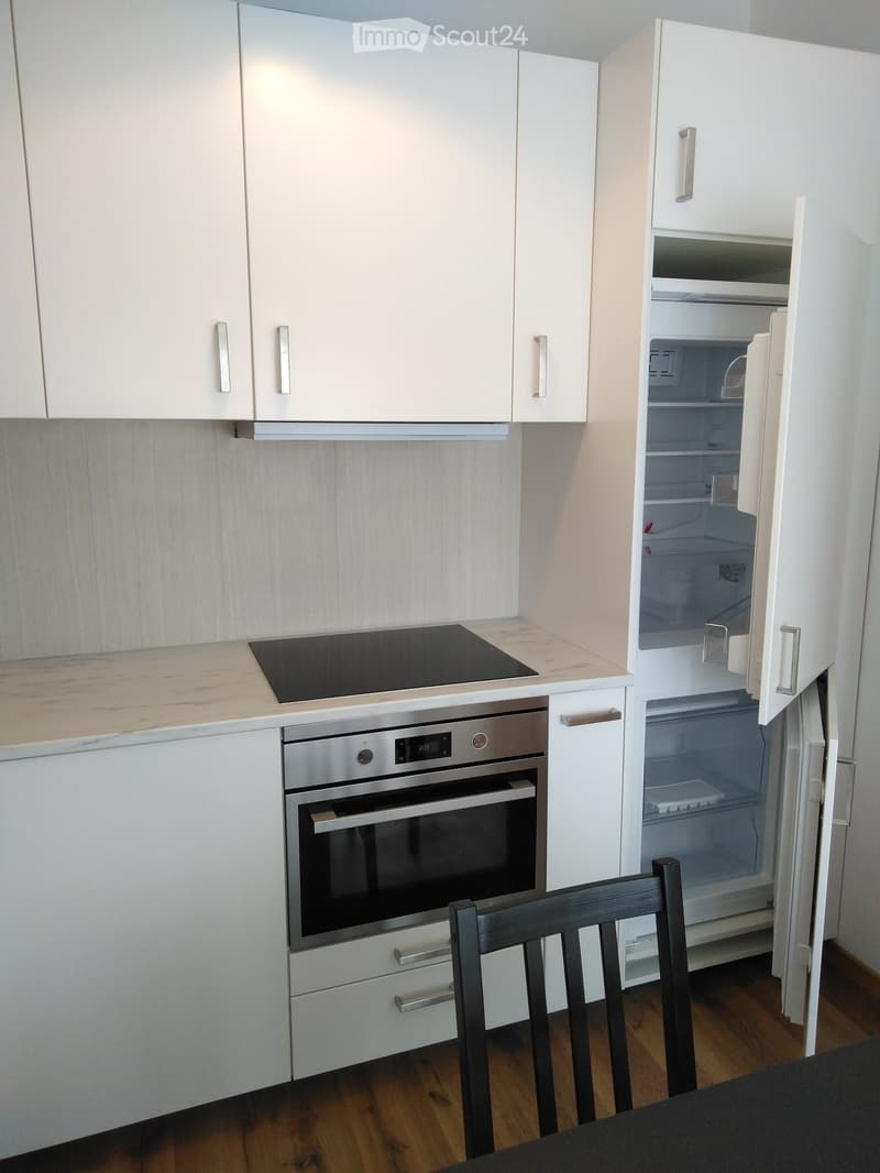 Modern kitchen with white cabinets, stainless steel oven, induction cooktop, and open refrigerator. Wooden floor and a single chair visible.