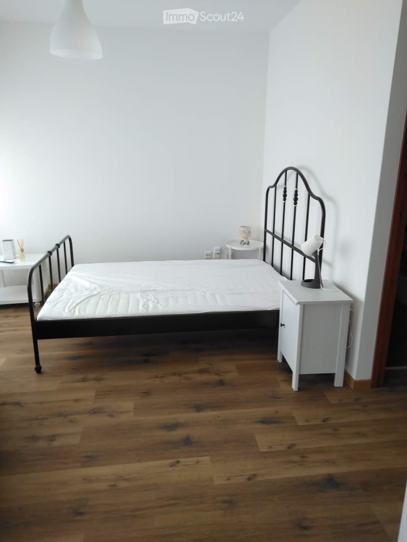 Minimalist bedroom with wooden flooring, a single bed with a white mattress, a nightstand with a lamp, and a small table with a lamp.