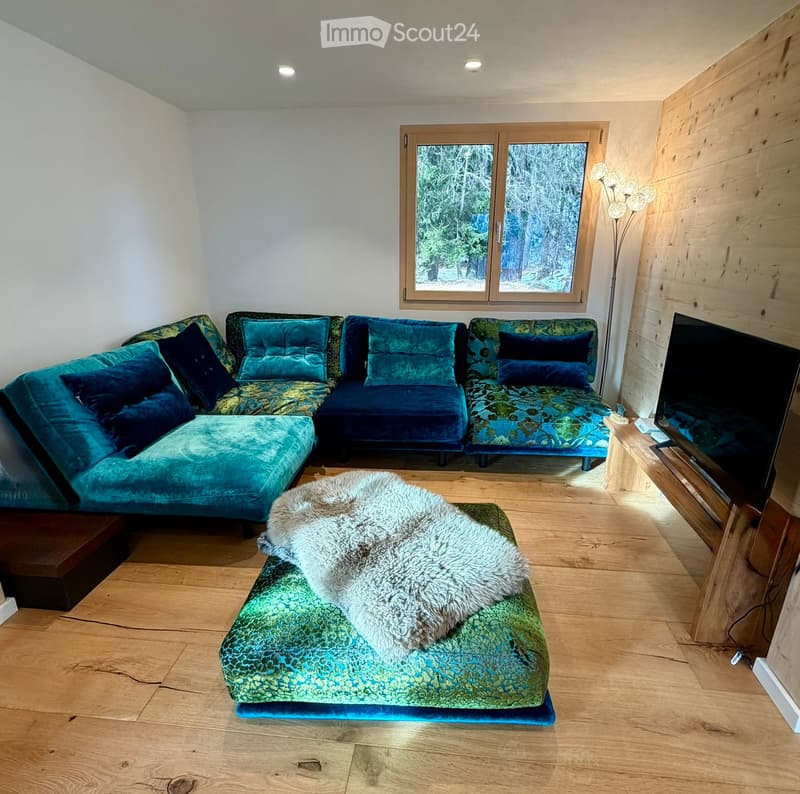 Living room: wooden floor, teal L-shaped sofa, ottoman with a rug, wooden TV stand, white walls, window, lamp