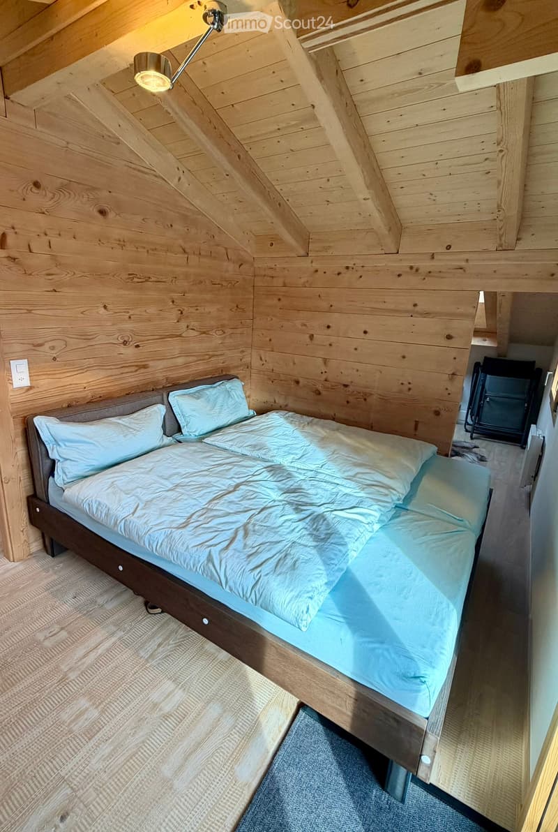 double bed with blue sheets, pillows, wooden walls, wooden floor, ceiling light, power outlet, chair in corner