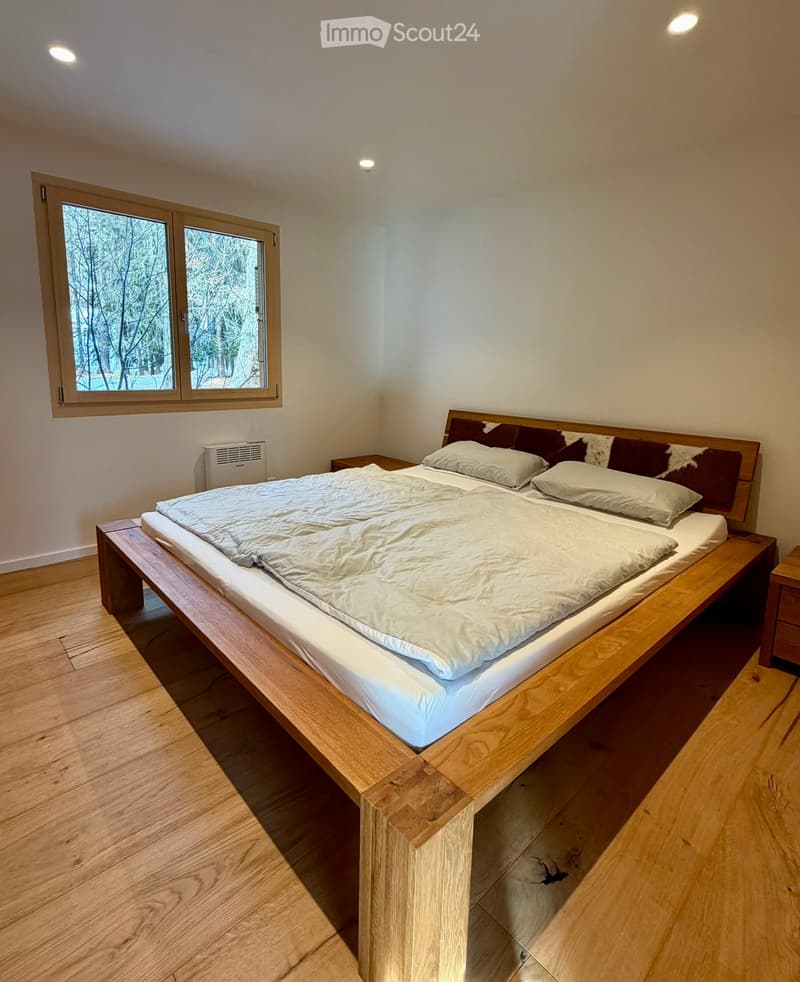 Bedroom with wooden bed, white bedding, pillows with cowhide trim, wooden nightstands, air conditioning, wooden flooring, and window overlooking trees.