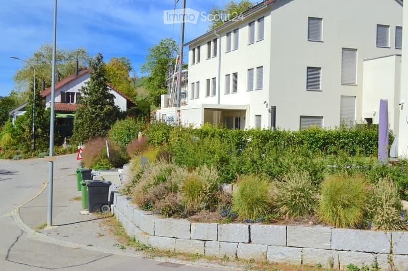 A modern, multi-story residential building with white walls and multiple windows, surrounded by a landscaped garden area with shrubs and plants.