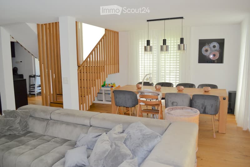 Open-plan living and dining area with a grey sectional sofa, wooden dining table with chairs, a small cat basket, and a staircase leading to another level.