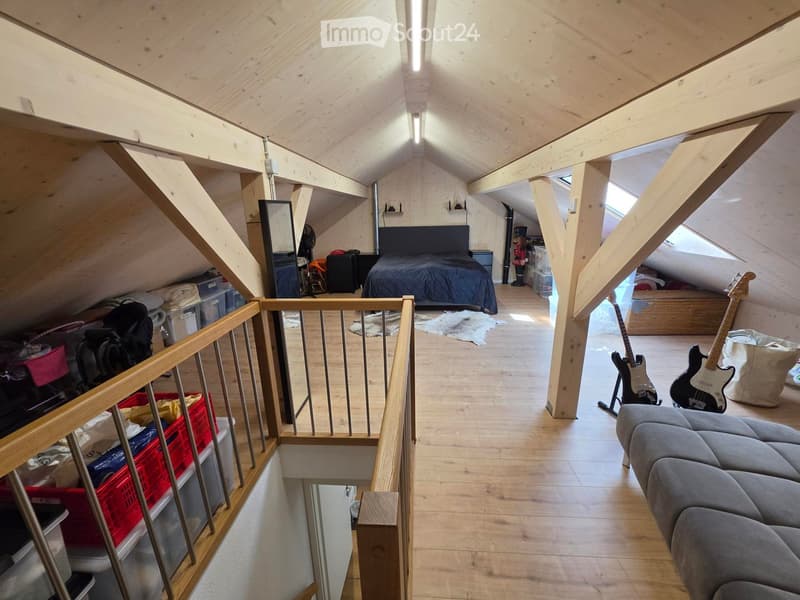 Loft-style room with wooden beams, slanted ceiling, and skylights. Features a grey sectional sofa, guitars on stands, a bed with dark bedding, and storage bins along the walls.