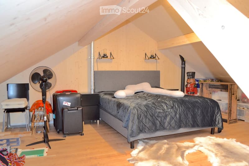 Cozy attic bedroom with wooden floors, slanted ceilings, and modern furnishings including a bed, nightstand, fan, chair, suitcase, and decorative items.