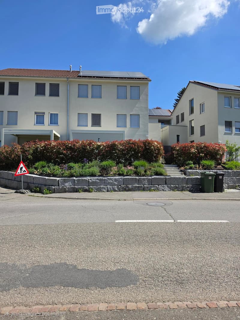 Two-story house with solar panels on the roof, multiple windows, and a landscaped front yard with bushes and a stone border.