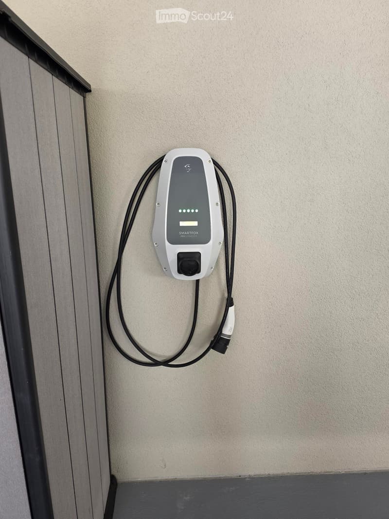 A white and grey electric vehicle charging station mounted on a beige wall, with a black cable extending from it.