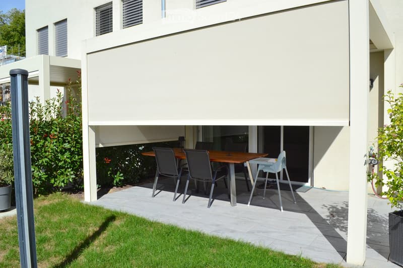 Outdoor patio with a wooden table, four chairs, and a baby chair, surrounded by a modern house with large windows and a retractable awning.