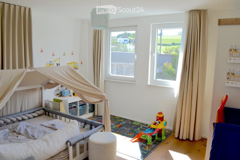 Brightly lit children's bedroom with a raised bed, canopy, colorful play area, and large windows overlooking a green landscape.