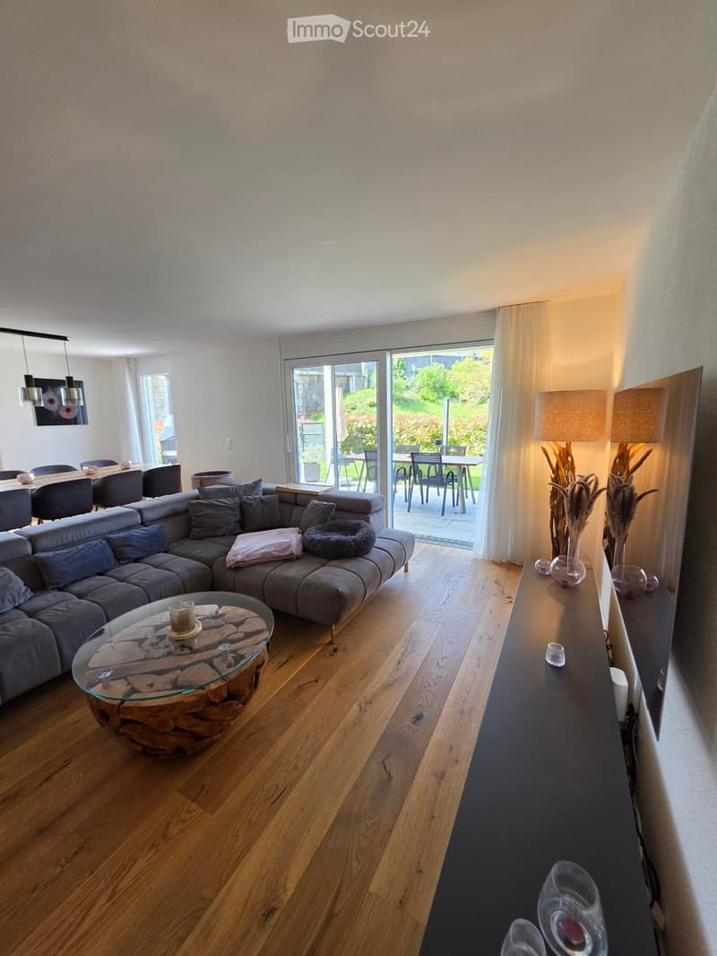 Modern living room with a grey sectional sofa, glass coffee table, dining area with chairs, and decorative lamps. Large sliding glass doors lead to a patio with outdoor furniture.
