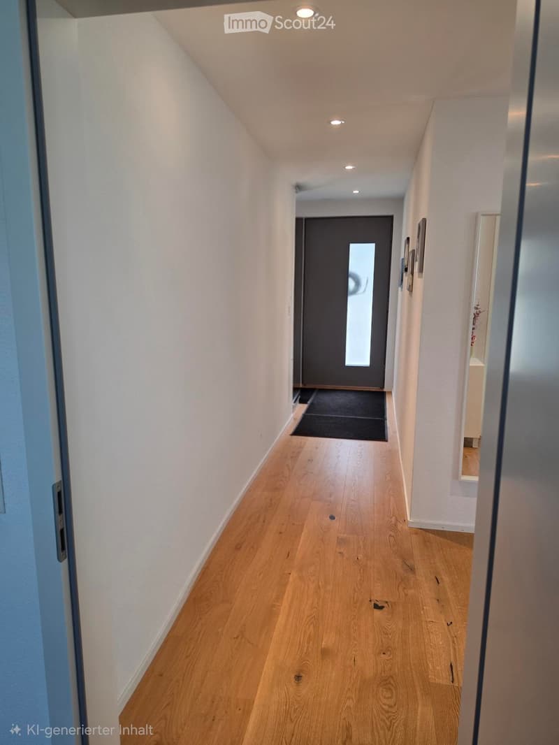 A clean, well-lit hallway with light wooden flooring, white walls, and recessed ceiling lights. A dark door is visible at the end, with a small black rug placed in front of it. The hallway is empty, with no furniture or decorations apart from a small fram