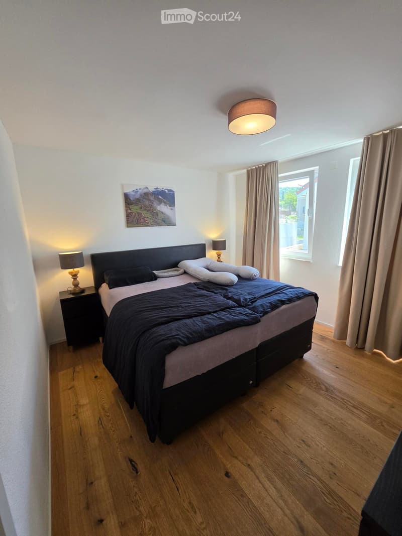 Modern bedroom with wooden flooring, large bed with dark bedding, nightstands with lamps, and a large window with curtains.