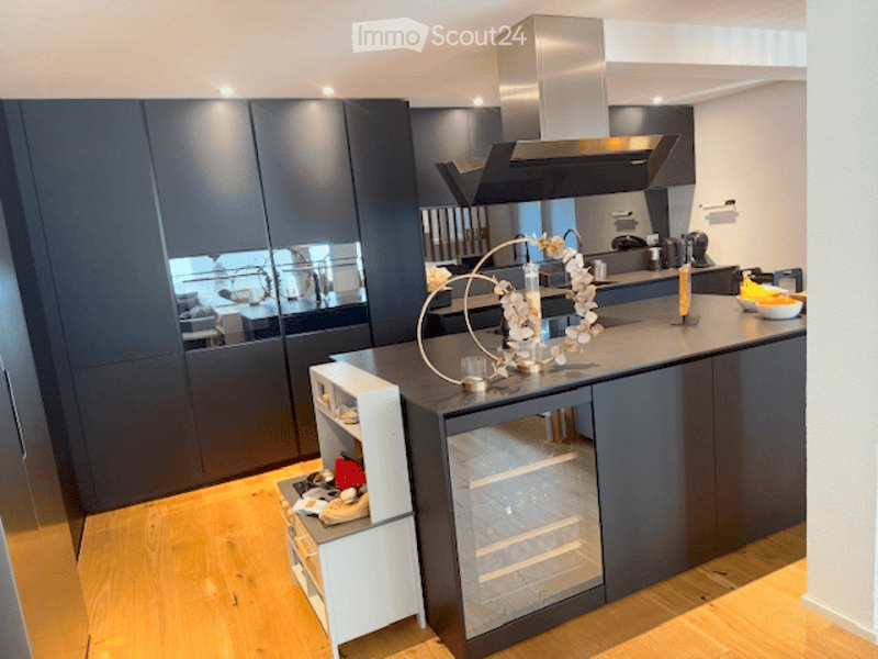 Modern kitchen with dark grey cabinets, a central island, and a stainless steel range hood. The island features a decorative arrangement and a bowl of fruit. A small white refrigerator is visible, along with a sink and faucet.