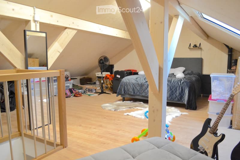Bright attic bedroom with wooden floors, slanted ceilings, and large windows. Contains a double bed, a guitar, a fan, a mirror, a rug, and a toy.