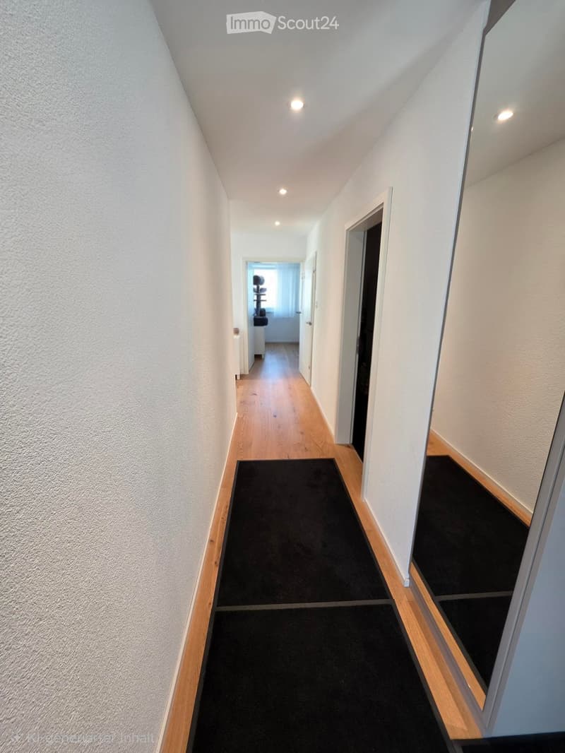 A narrow hallway with light wooden flooring, white walls, and black runner rugs. The ceiling has recessed lighting, and there are several doors leading to other rooms.