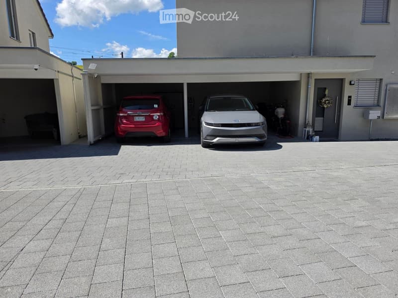 Garage with two cars parked inside, attached to a modern house with a paved driveway.